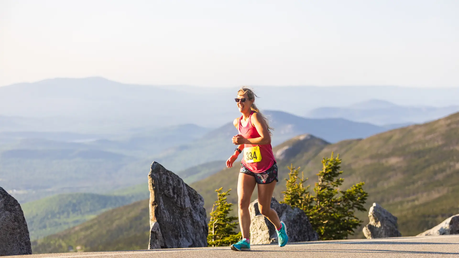 A female runner wearing a tank top, shorts, and sunglasses smiles while running during the Whiteface Mountain Uphill Race.