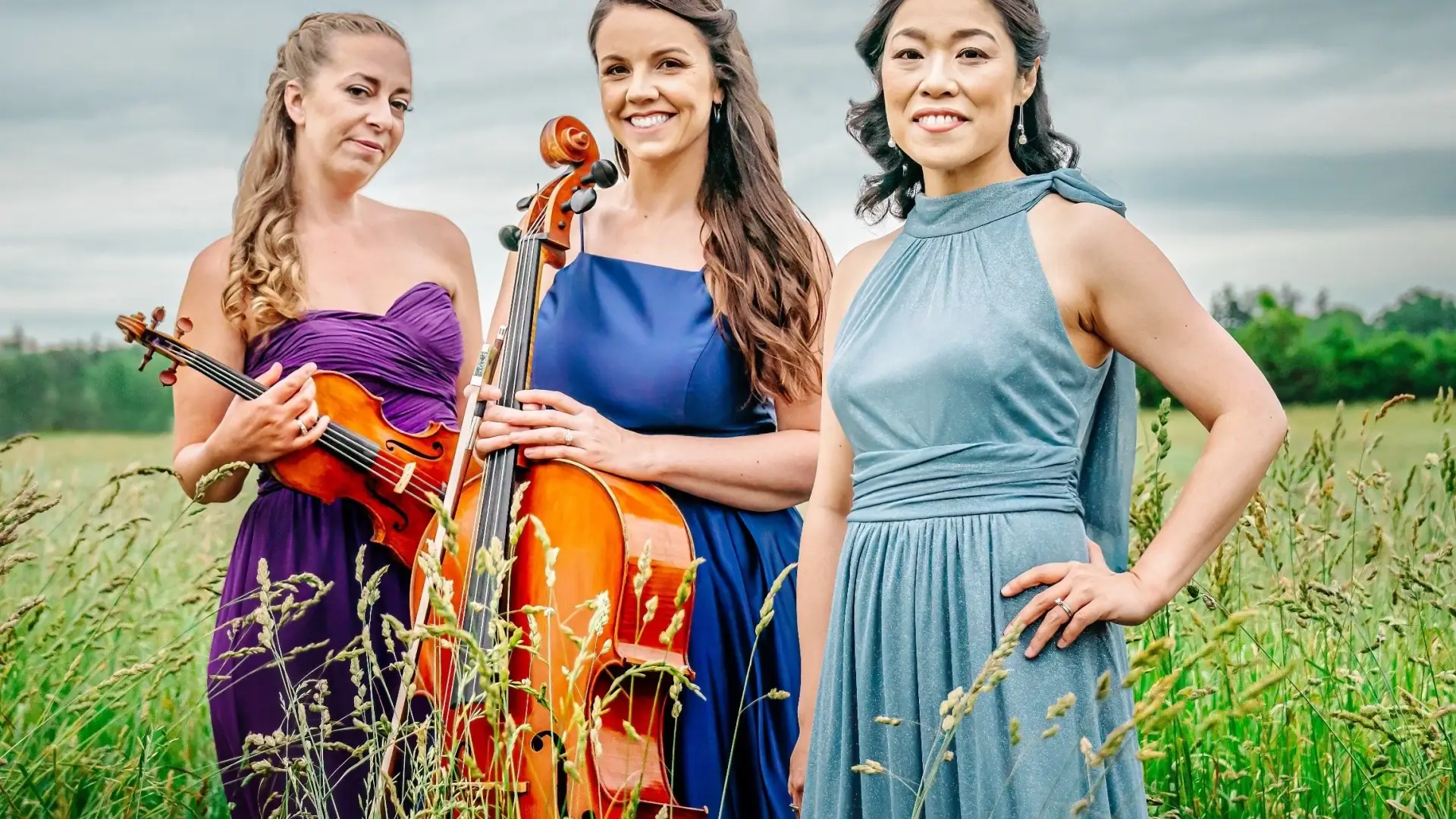 Three women in elegant dresses stand in a field. One holds a cello, another a violin.