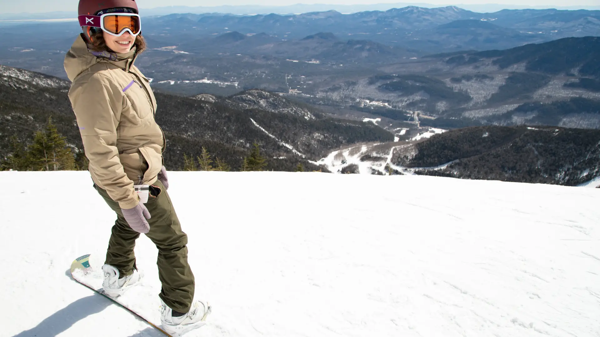 A smiling snowboarder at the top of a snowy run overlooking the main lodge of Whiteface Mountain.