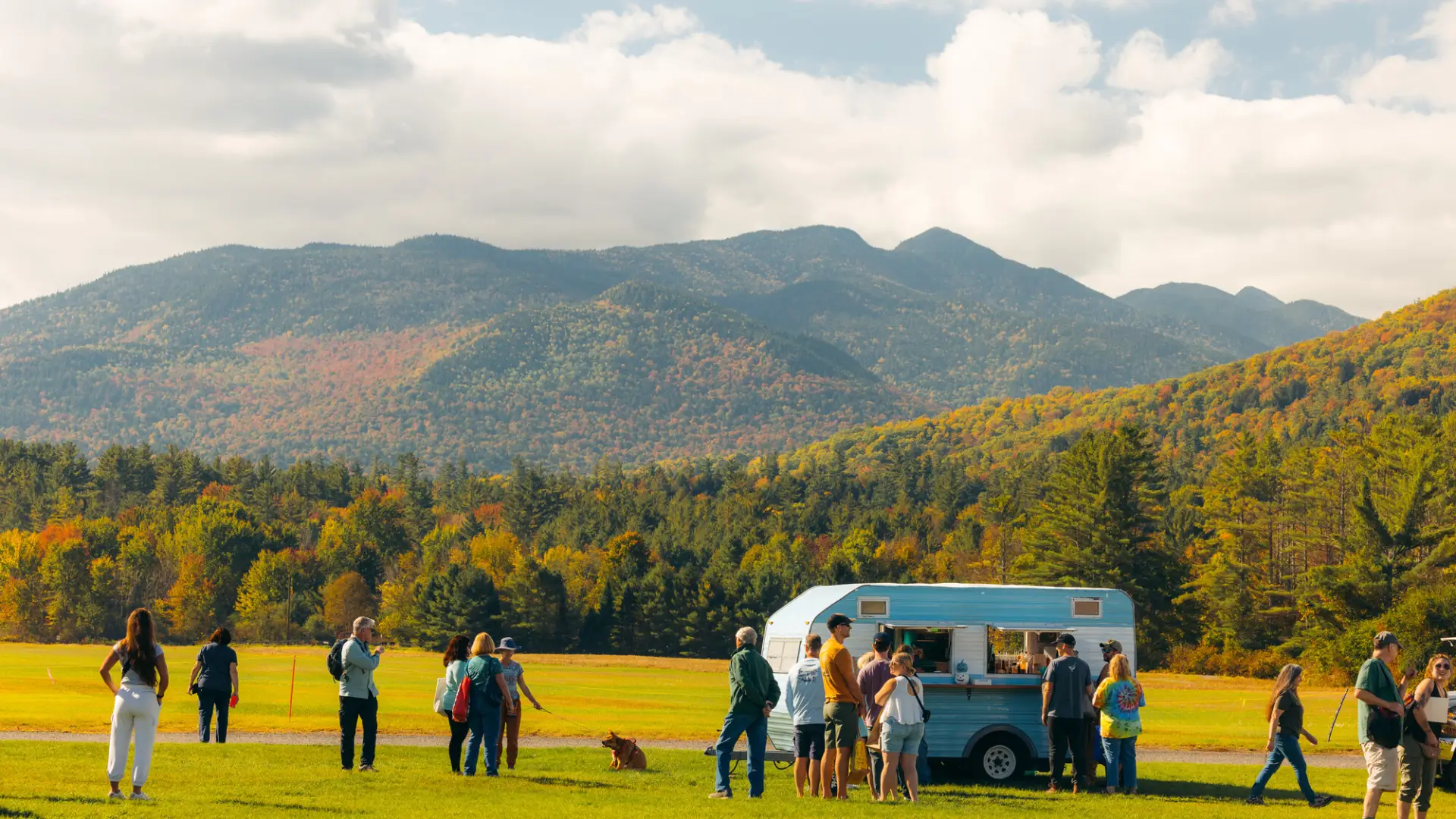 People stand around blue and white striped camper trailer in field with mountains in the background showing fall colors