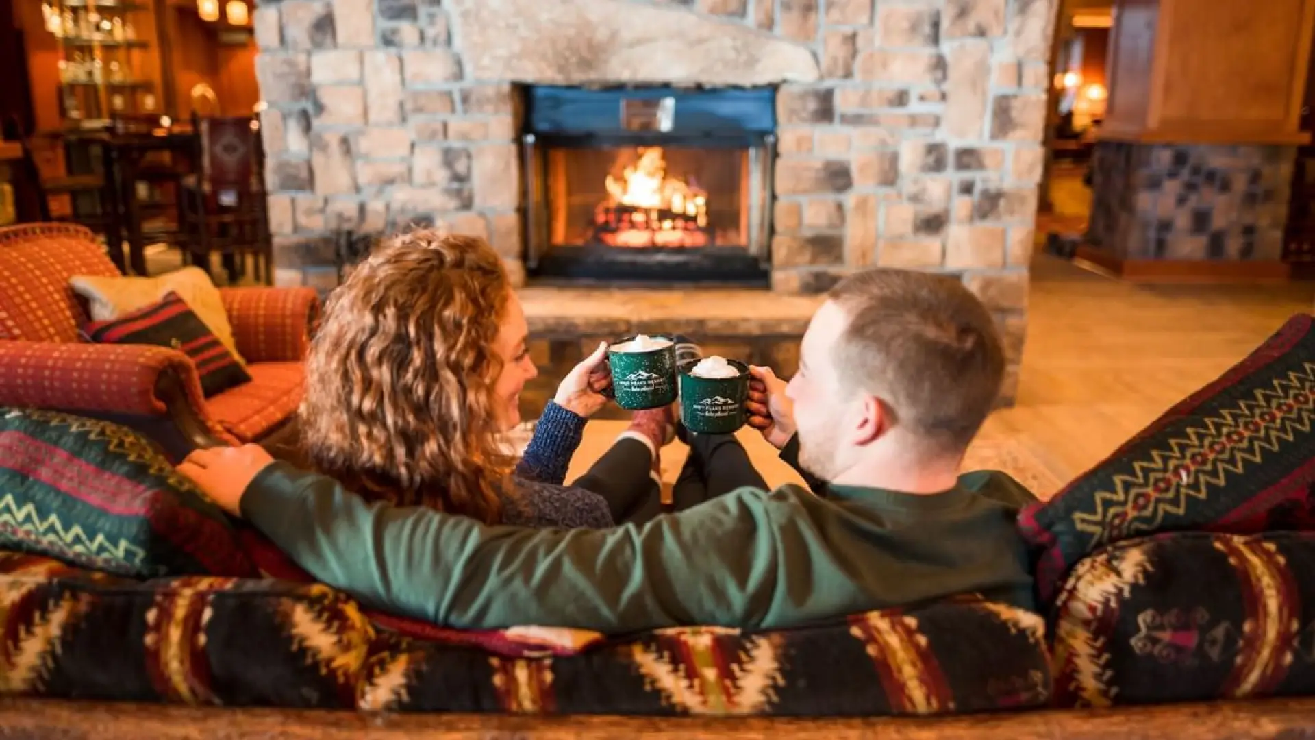A couple sits on a log couch in front of a large stone fireplace with cocoas