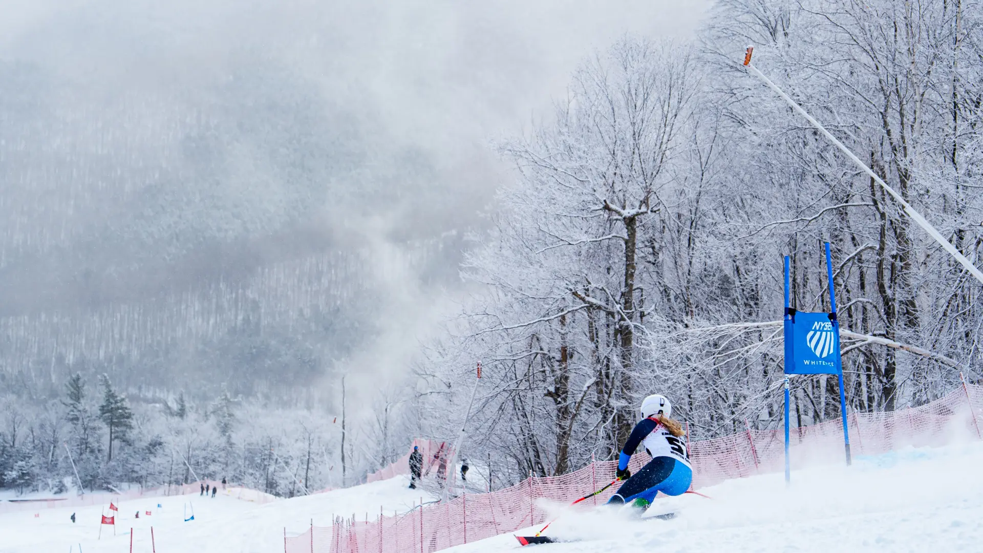 An alpine ski racer in a blue and black speed suit carves through a turn on a snow-covered mountain slope.