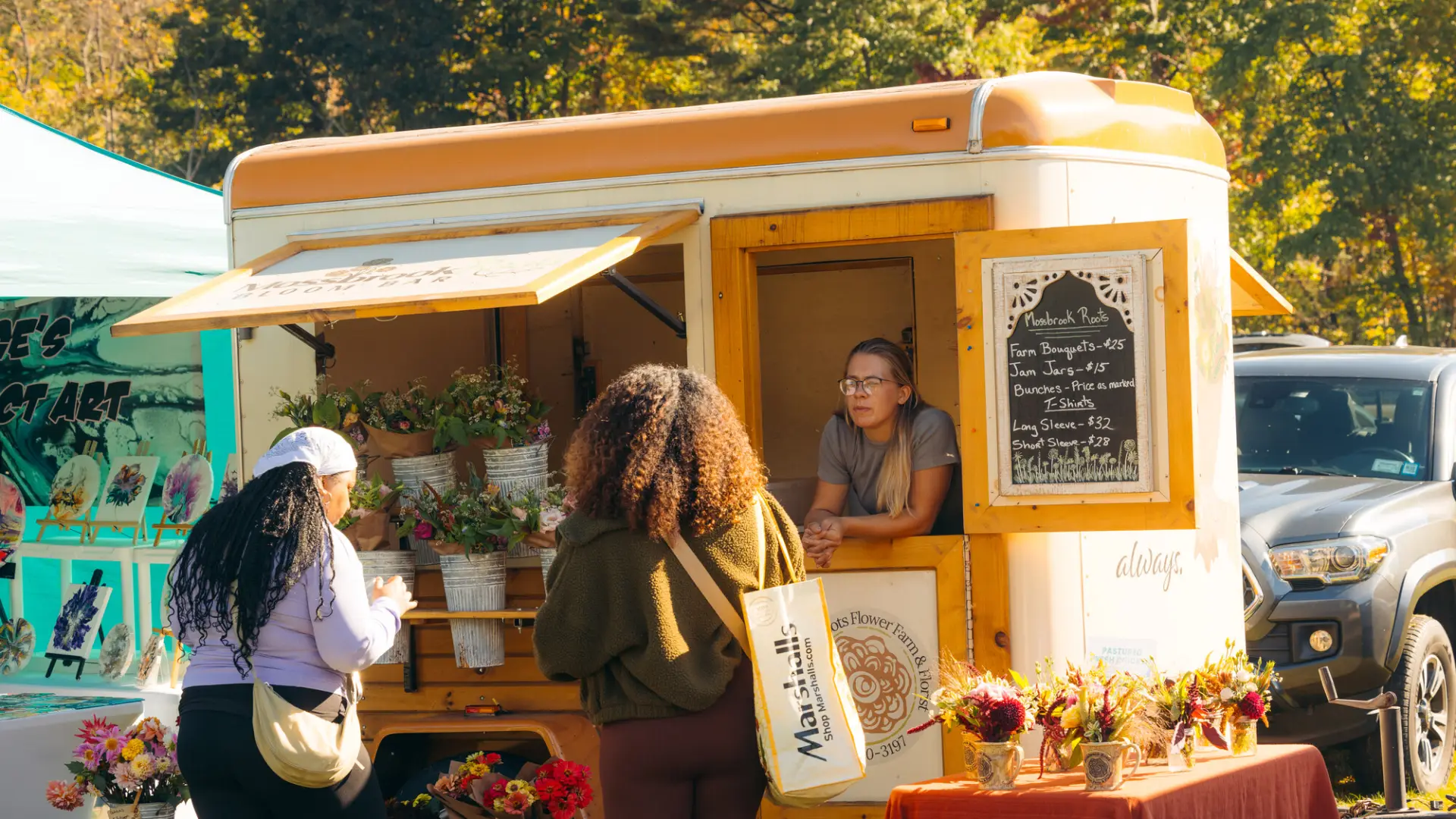 Two women browse flowers in metal buckets hung on side of camper trailer with woman leaning out window talking to customers