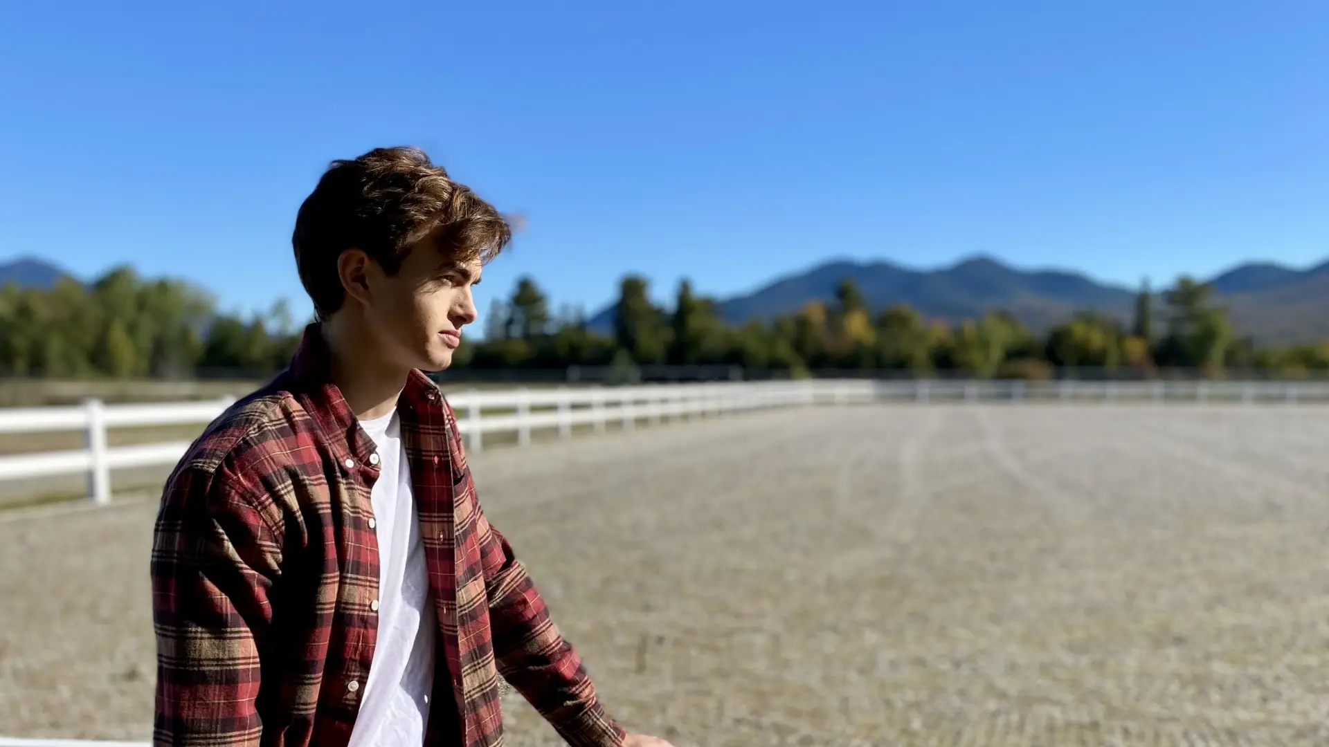 A young man in a flannel and jeans stands next to a white fence.