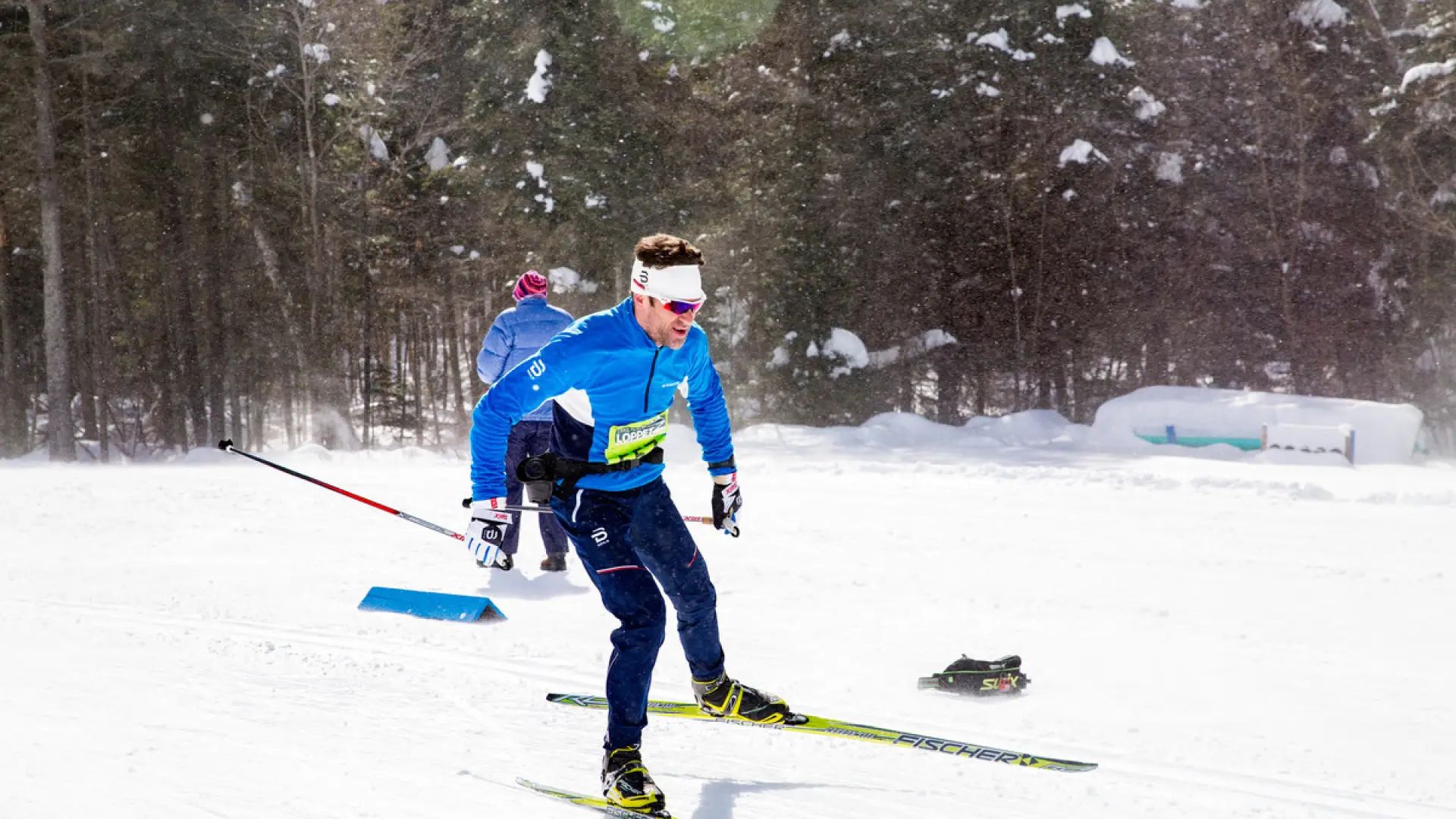 A skier in blue and black cross-country ski gear skis past a course worker during the Lake Placid Loppet race. The skier has a yellow race bib on their chest.