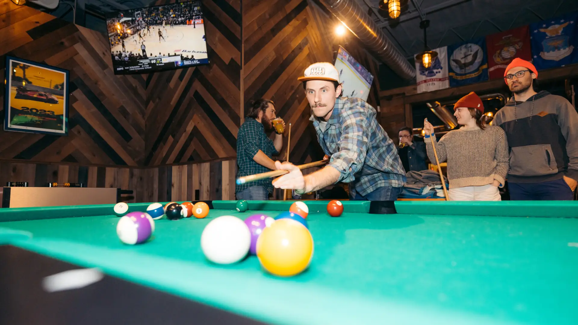 Young man with a mustache wearing flat brim hat lines up a shot at a pool table with friends behind him