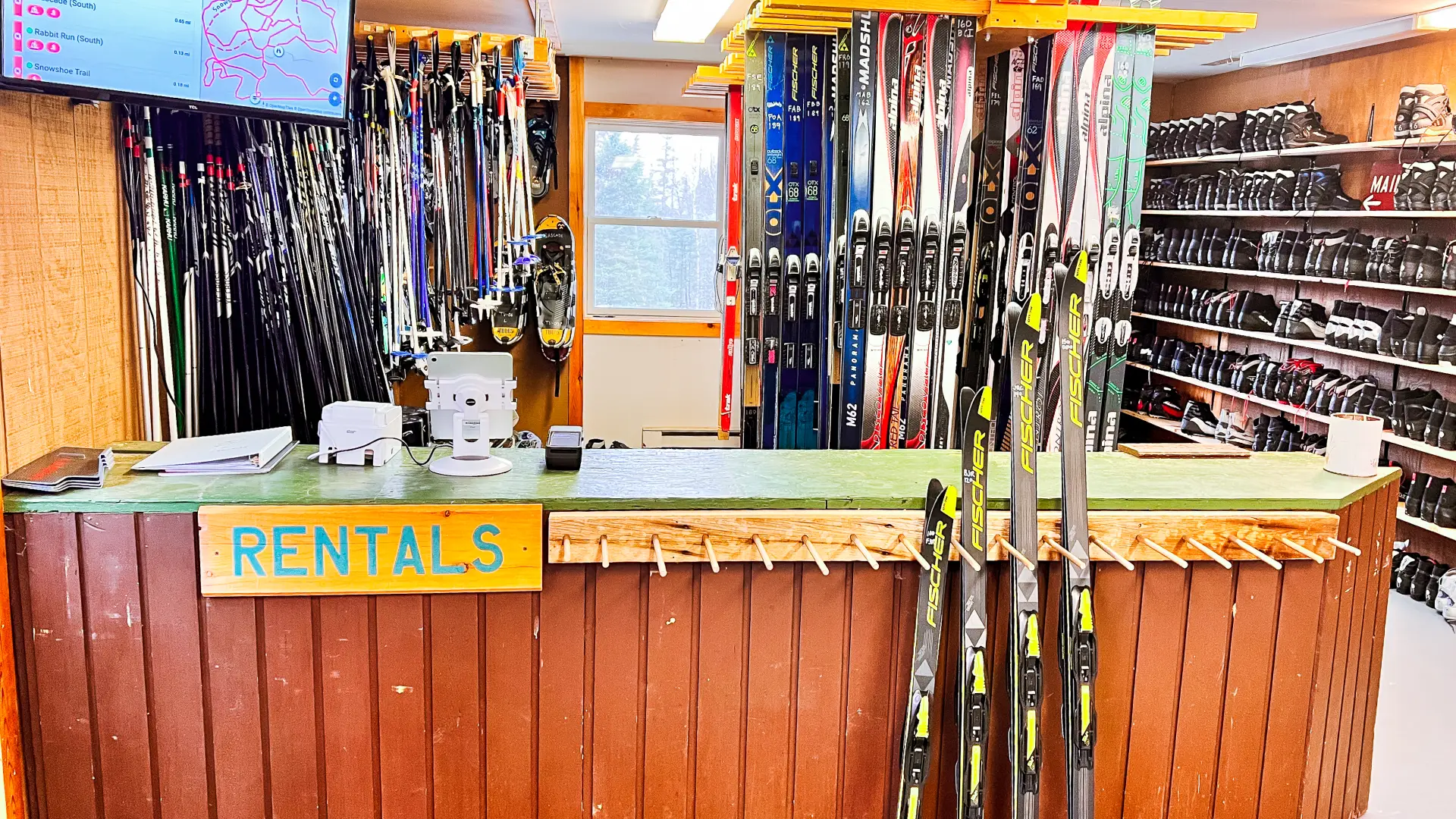 A full rental room with four pairs of skis set out for display. Boots, skis, and poles in the background for rent.