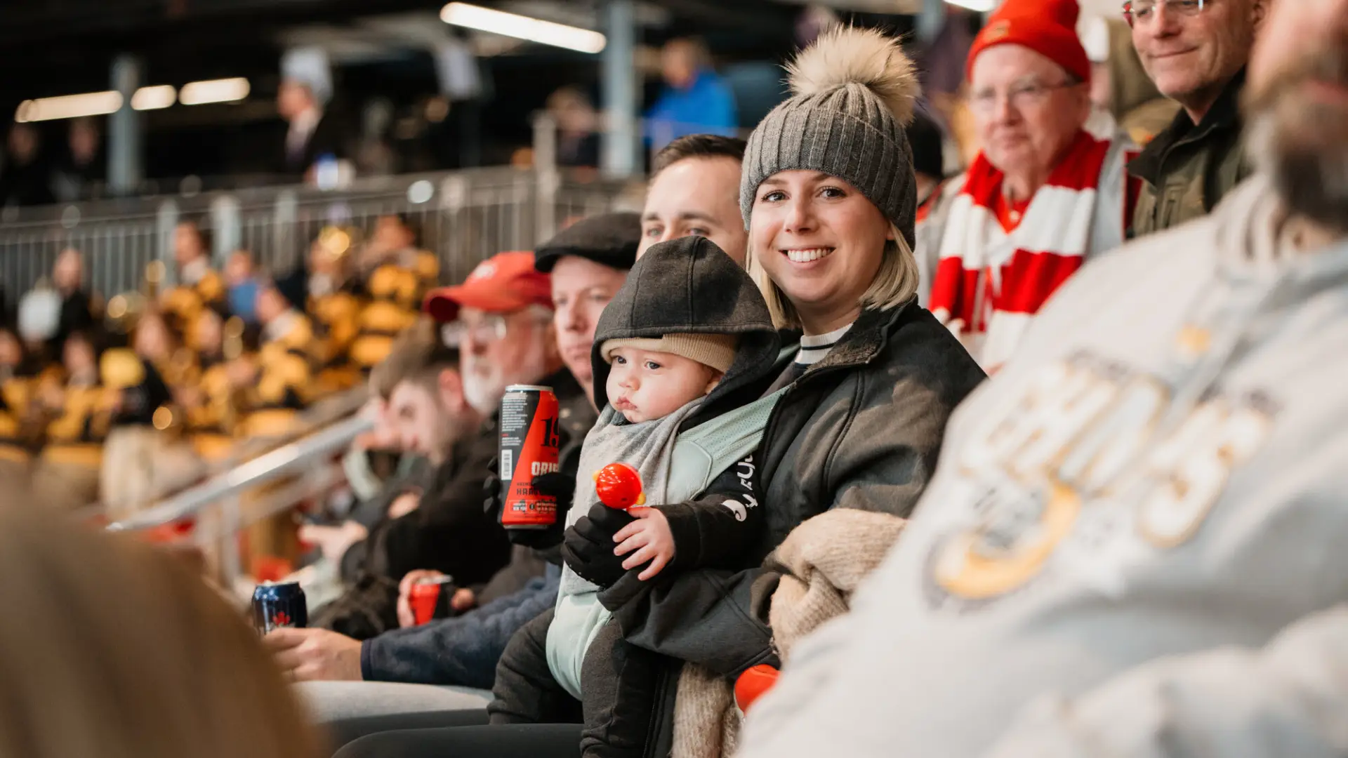 a woman holds her baby and drink while spectating a sporting event in an arena. 