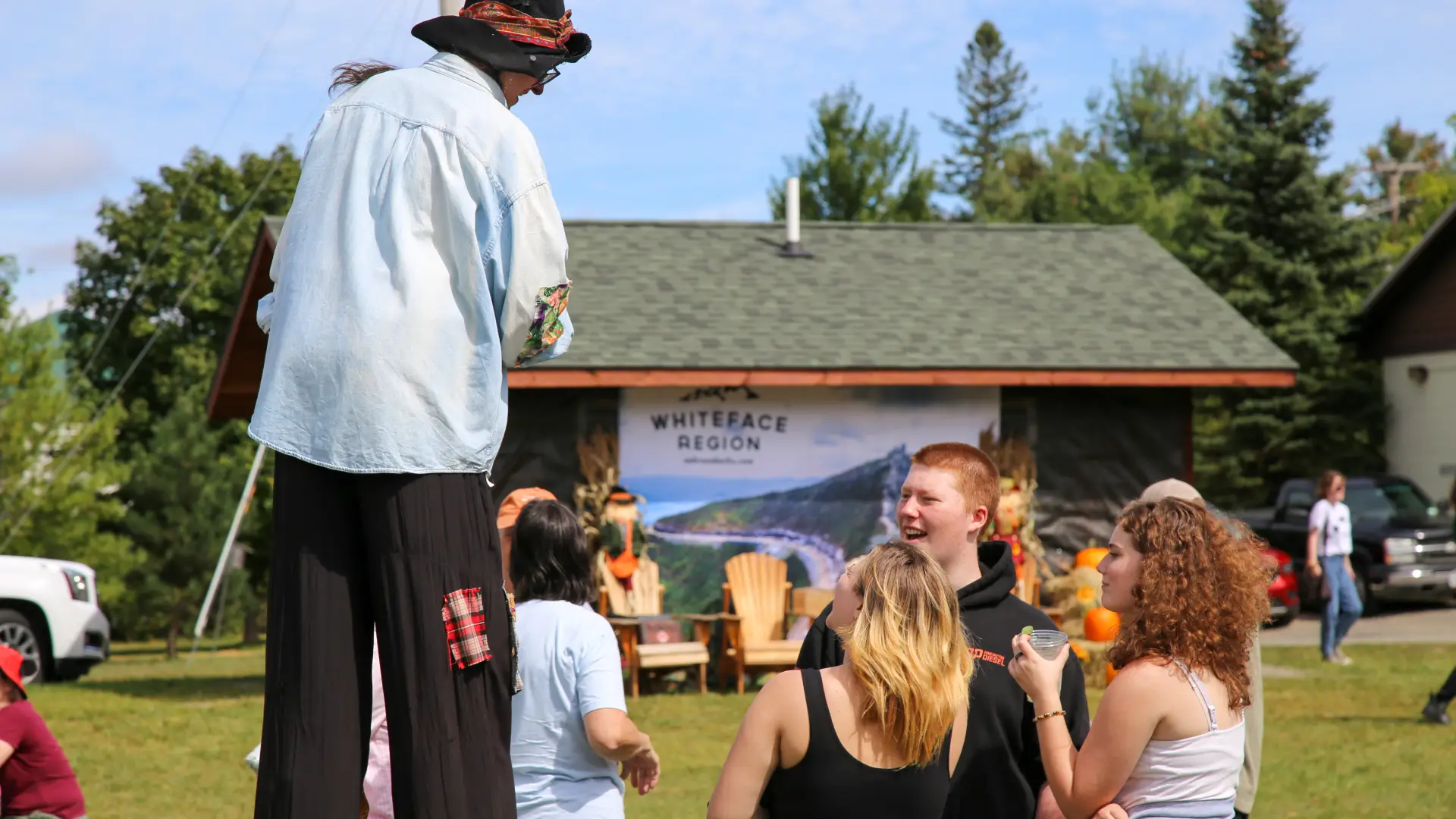 Collection of youths stand in a group talking to a woman dressed as a scarecrow on stilts