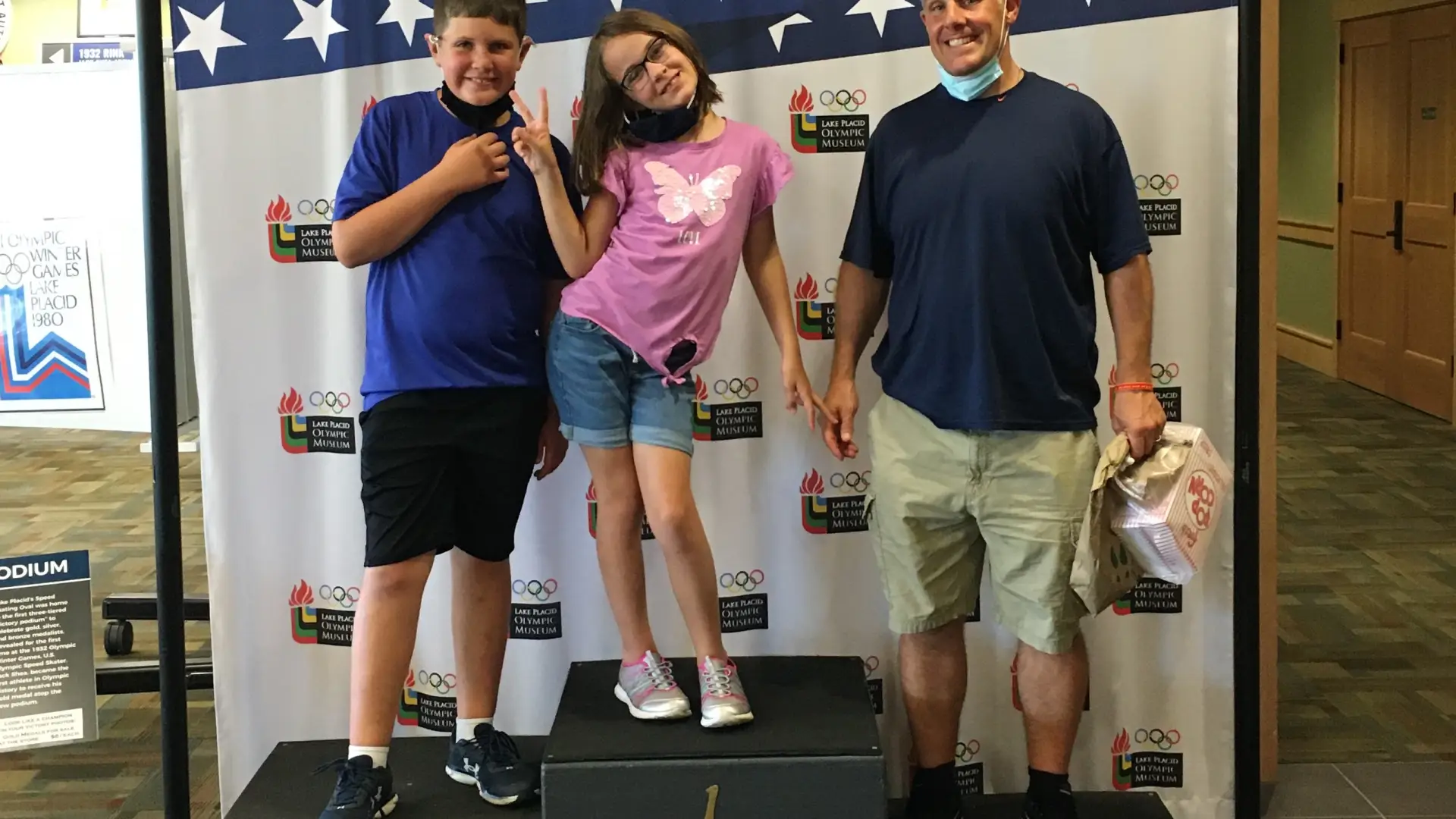 Two children and an adult male stand on a pretend Olympic medal stand in front of a stars and stripes backdrop.