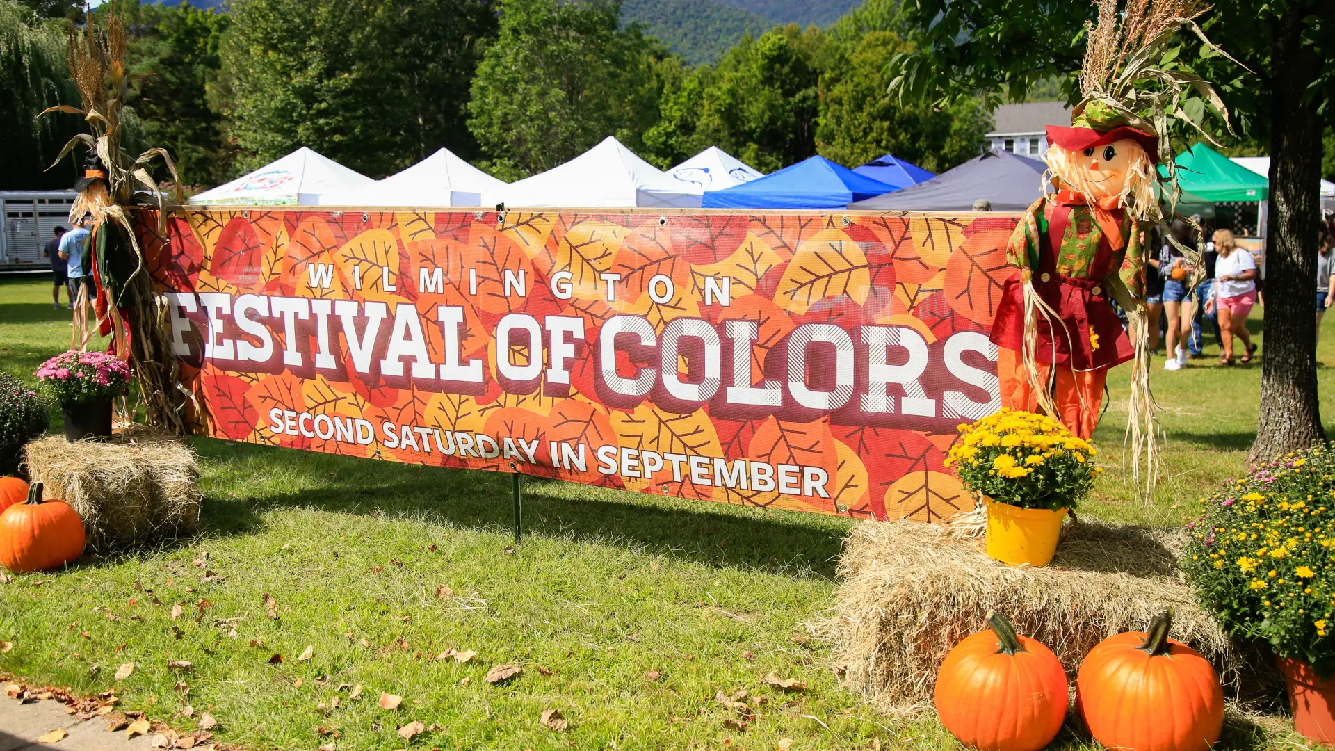 Large horizontal Festival of Colors banner in front of event bookended by hay bales, mums, pumpkins, and scarecrows