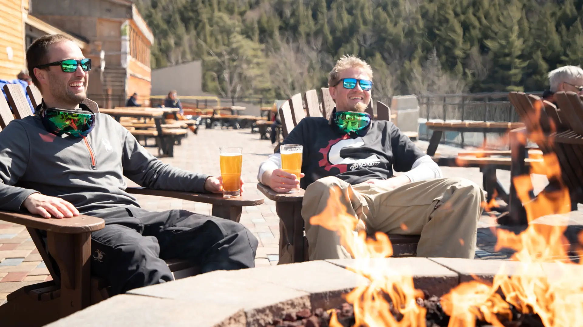 Two bros sit by a stone fire pit at a ski resort, laughing in Adirondack chairs with glasses of golden-colored beer.