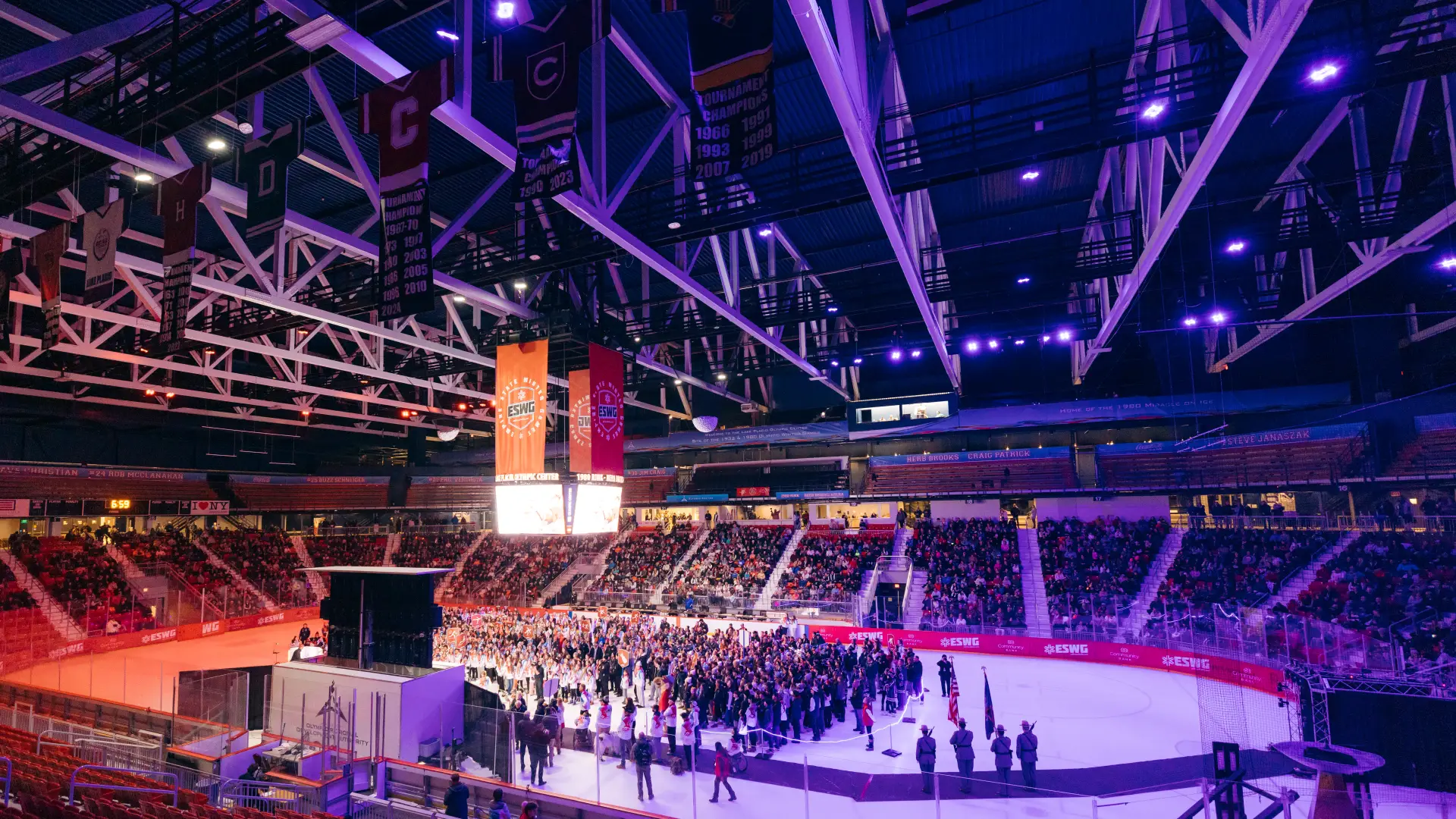 A wide-angle, high-angle shot captures a crowded ice arena during the 2025 Empire State Winter Games opening ceremony, featuring athletes gathered on the ice under purple stadium lighting and championship banners hanging from the rafters.