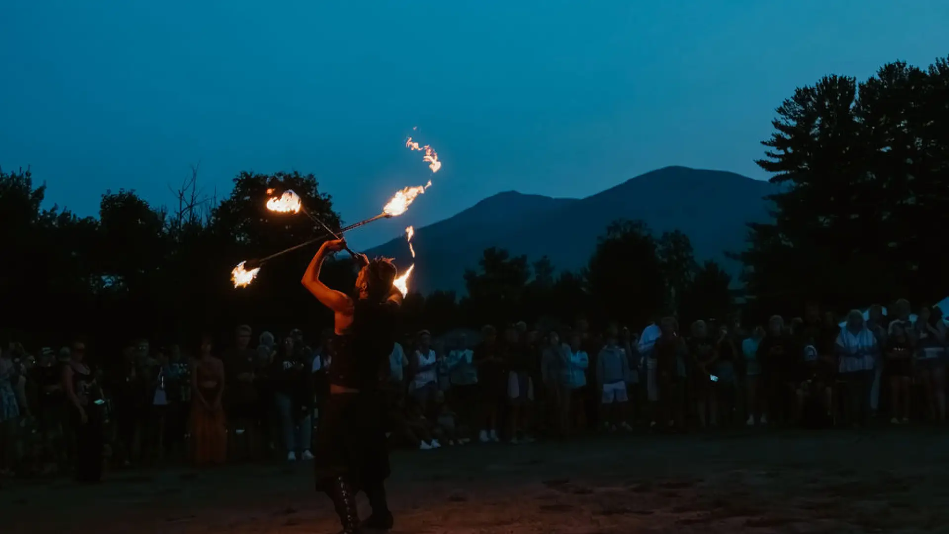 Fire dancer seen performing against backdrop of mountains and music festival at dusk
