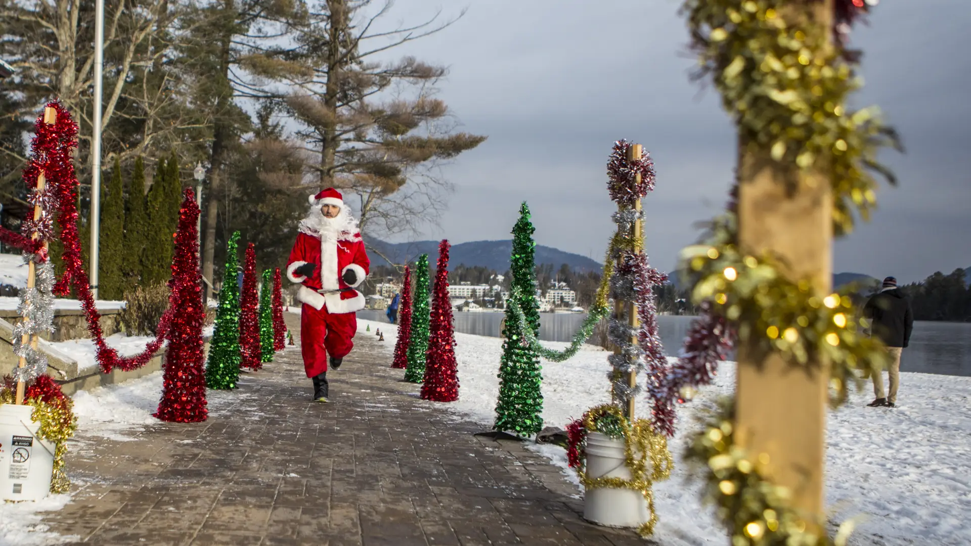 A person in a Santa costume happily jogs along a snow-covered path lined with festive, colorful tinsel decorations and small tree figures.