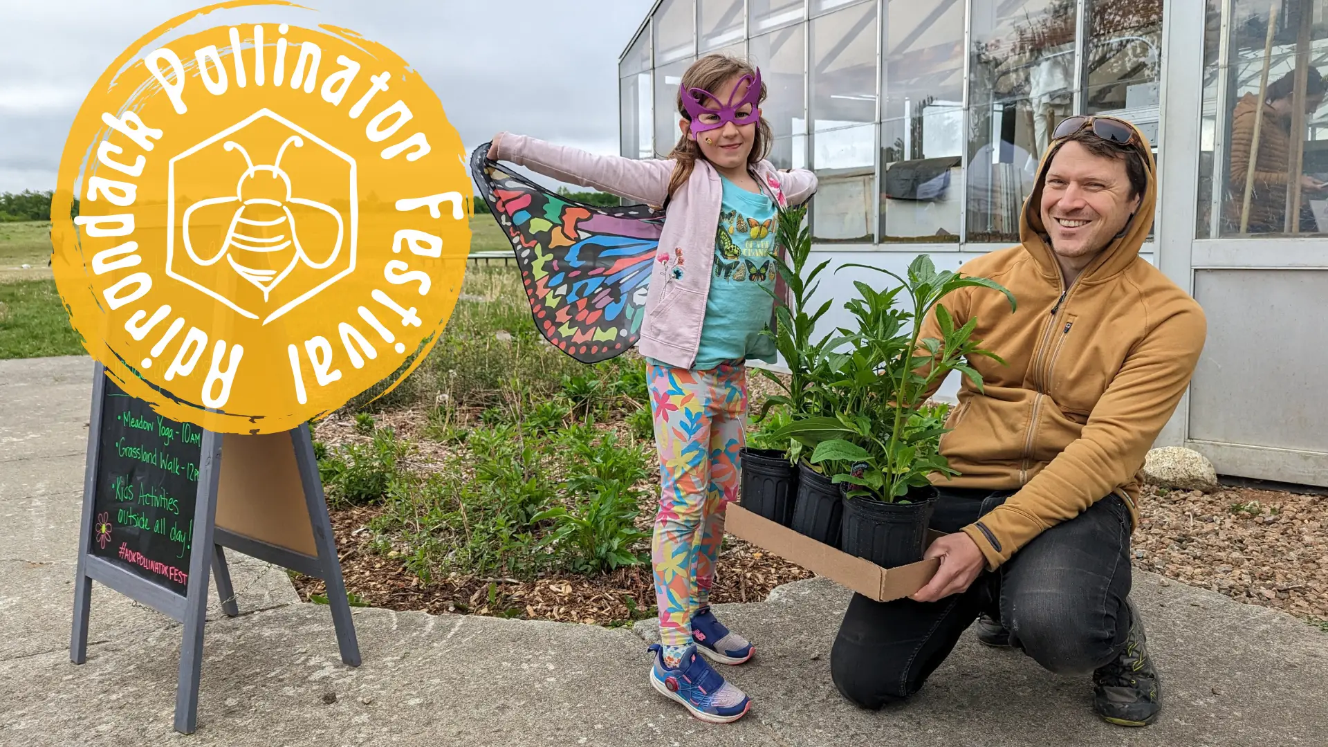 Man kneels holding flat of plants next to a young girl dressed in a butterfly costume in front of greenhouse