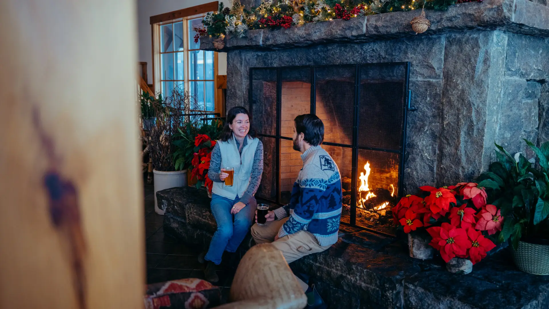 Couple sits side by side on the hearth of a stone fireplace with the fire behind them, each with a pint in their hand
