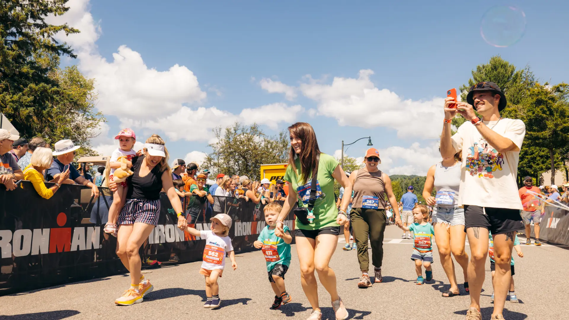 Collection of parents carrying some children and holding the hands of others walk on Ironkids race course between Ironman fencing lined with spectators