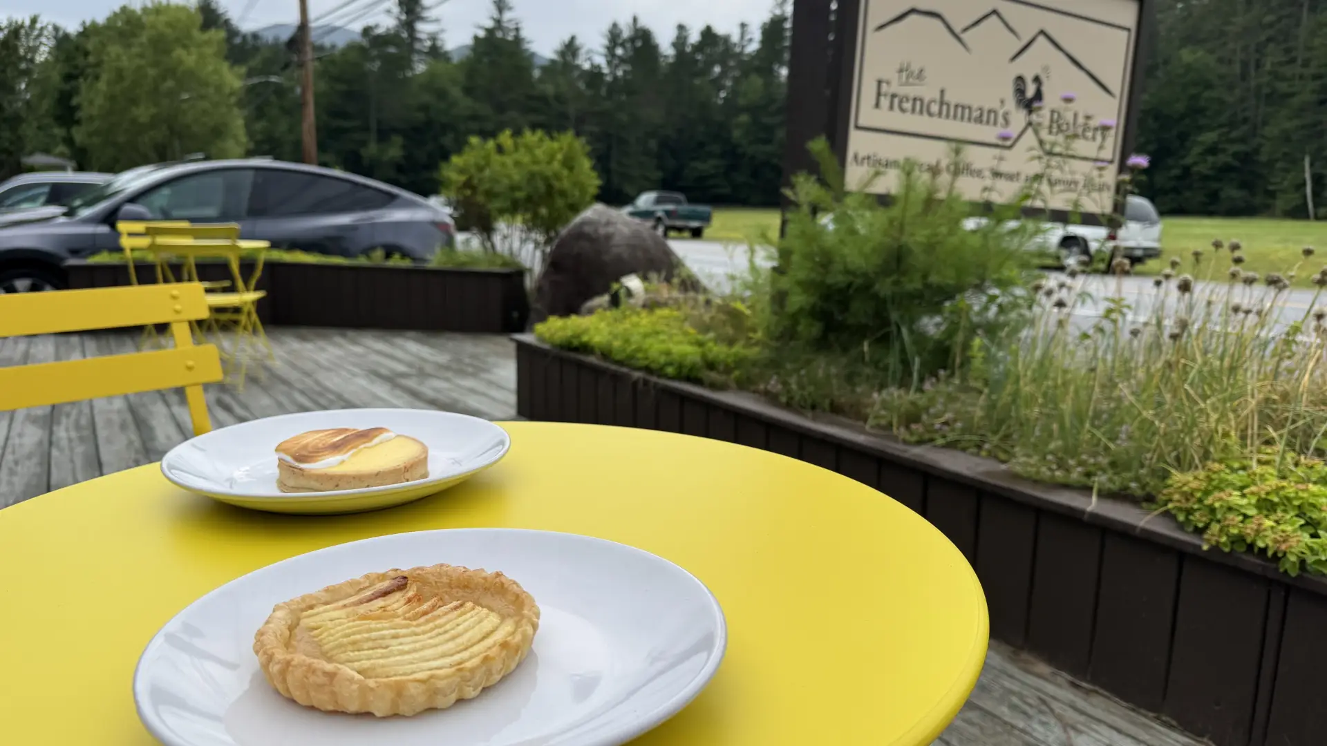 Two pastries sit on white plates on the yellow bistro tables on the wooden front deck of The Frenchman's Bakery.
