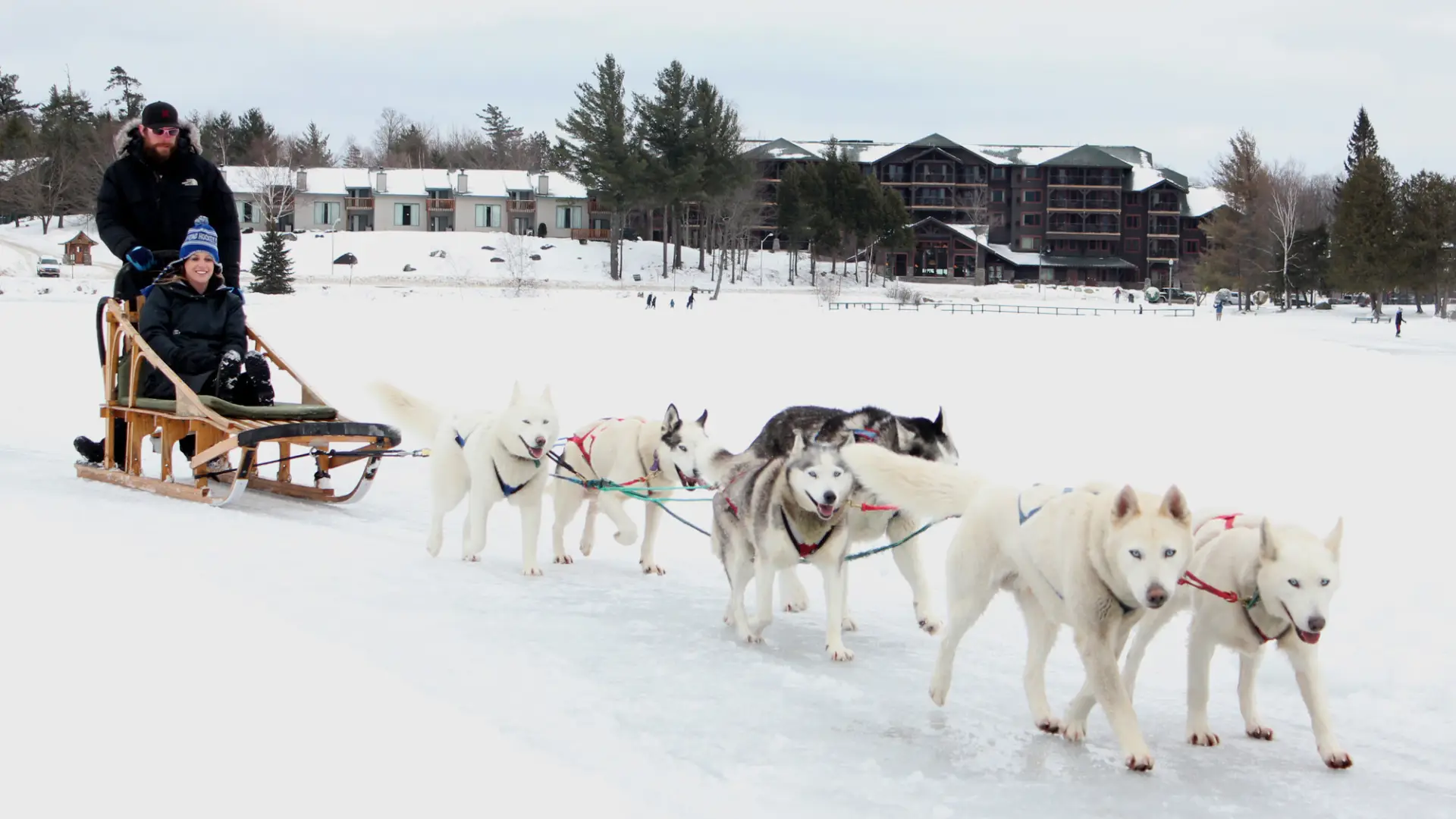 A dog team pulls a sled across a snowy field with a resort in the background.