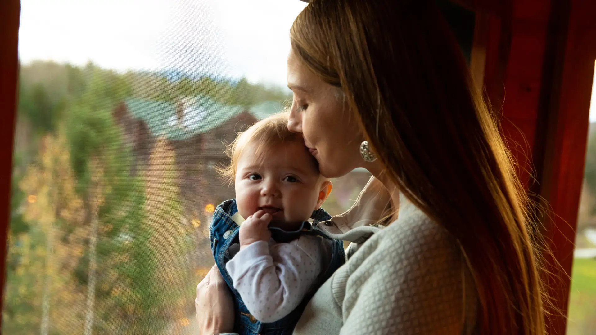 Woman kisses top of baby's head standing in large window overlooking trees and lodge-style resort