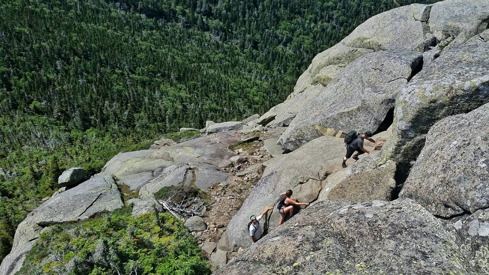 Three hikers scramble up vertical rocks on the side of a mountain.
