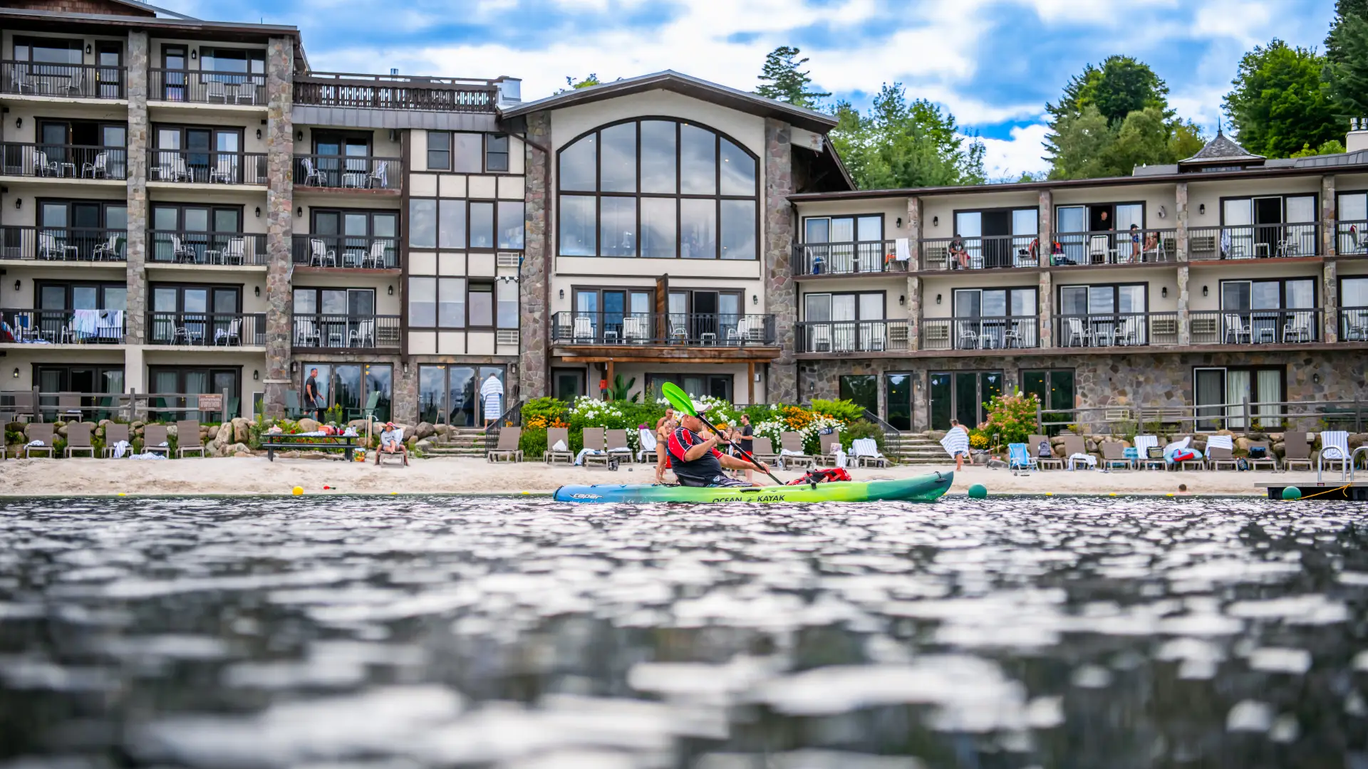 The mostly glass exterior of the Golden Arrow Resort, with the lake splashing and happy people on kayaks.