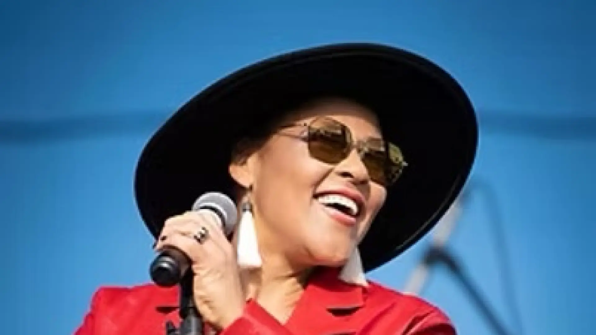 Dawn Tyler Watson on stage holding microphone waring red leather jacket, sunglasses, and black wide brim hat