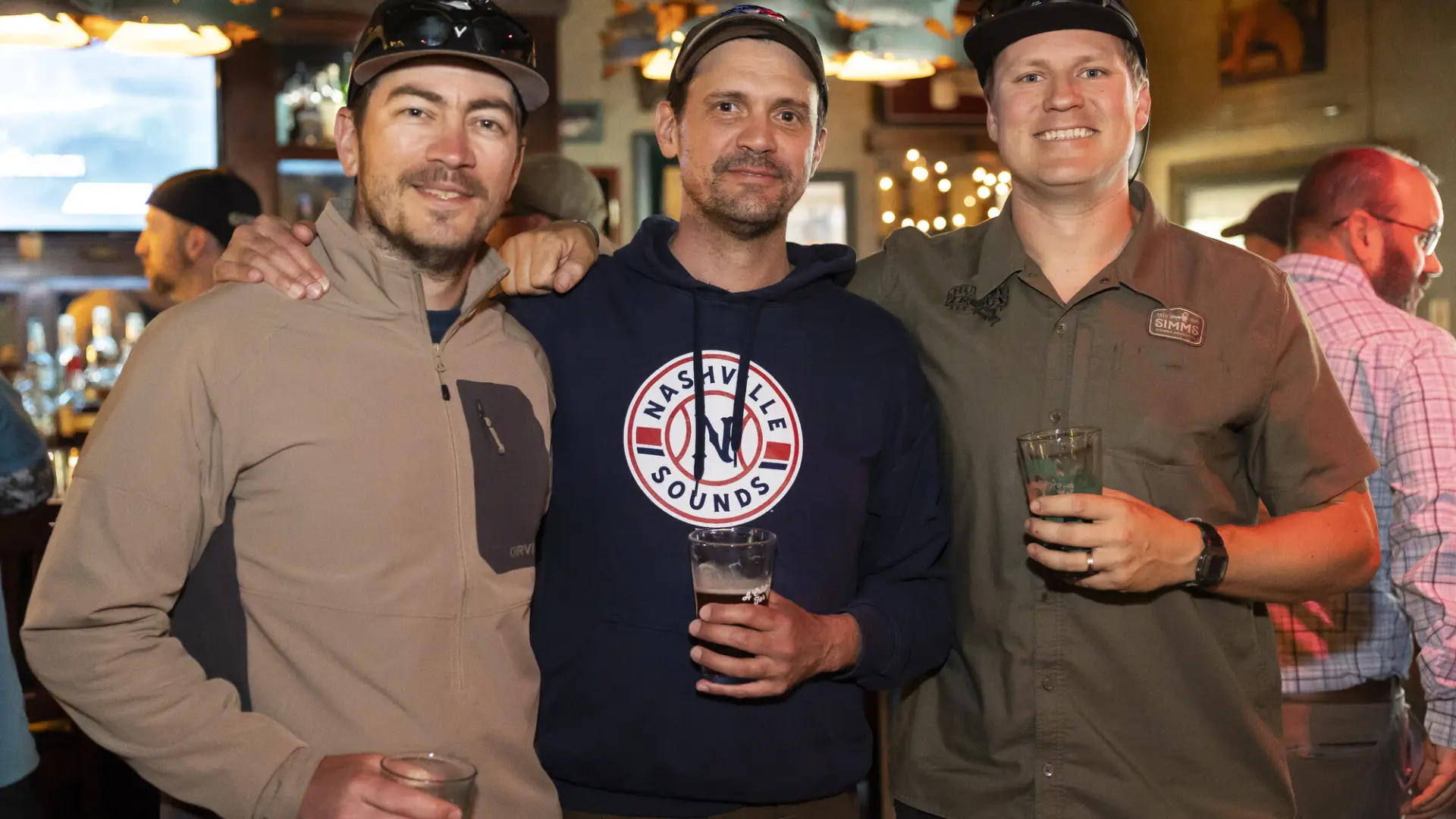 Three men stand together posing for photo with beer pints in hand at Ausable River Two-Fly Challenge event
