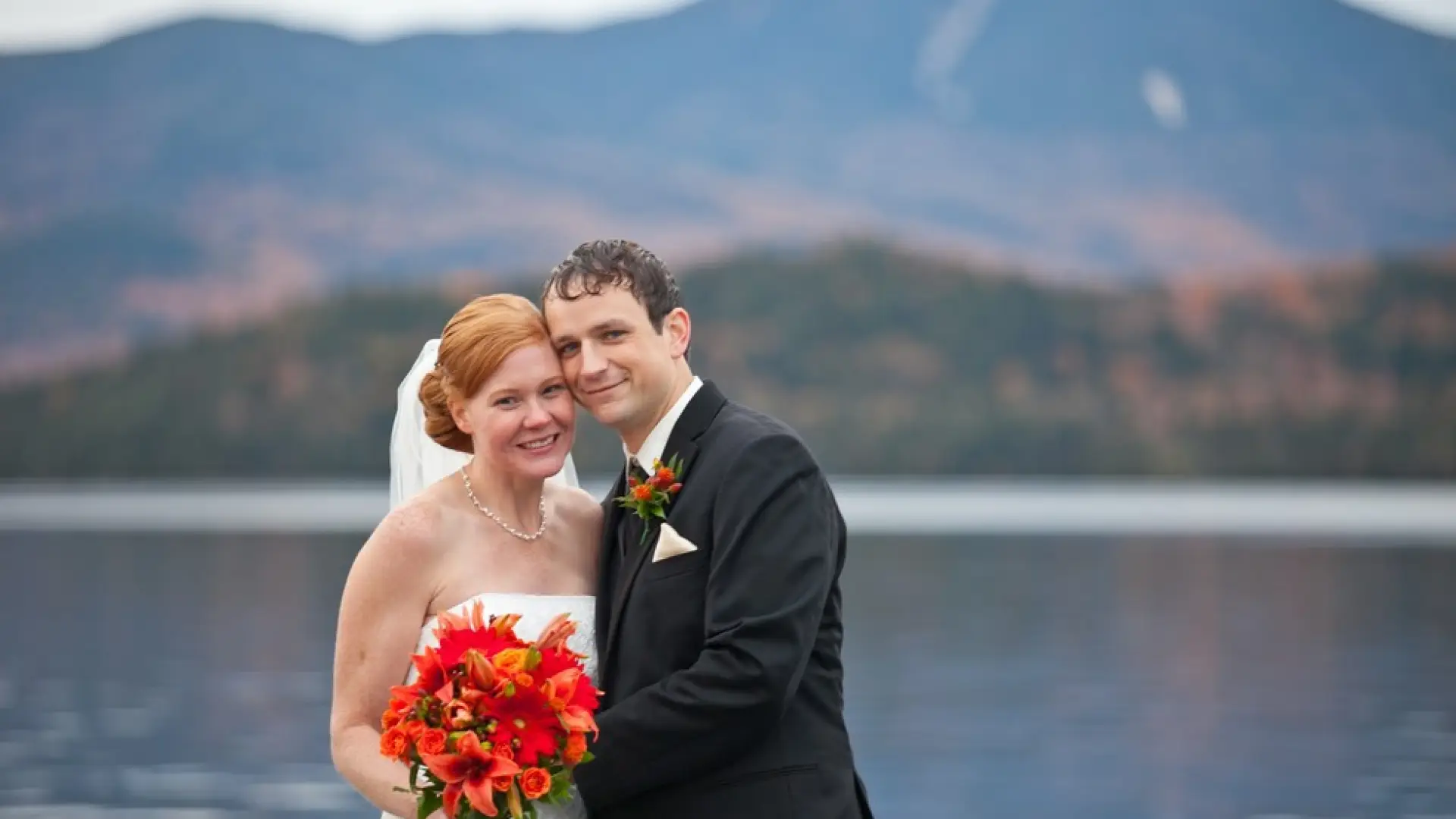 A happy couple smiles into the camera with the Mirror Lake and fall foliage covered mountains in the background