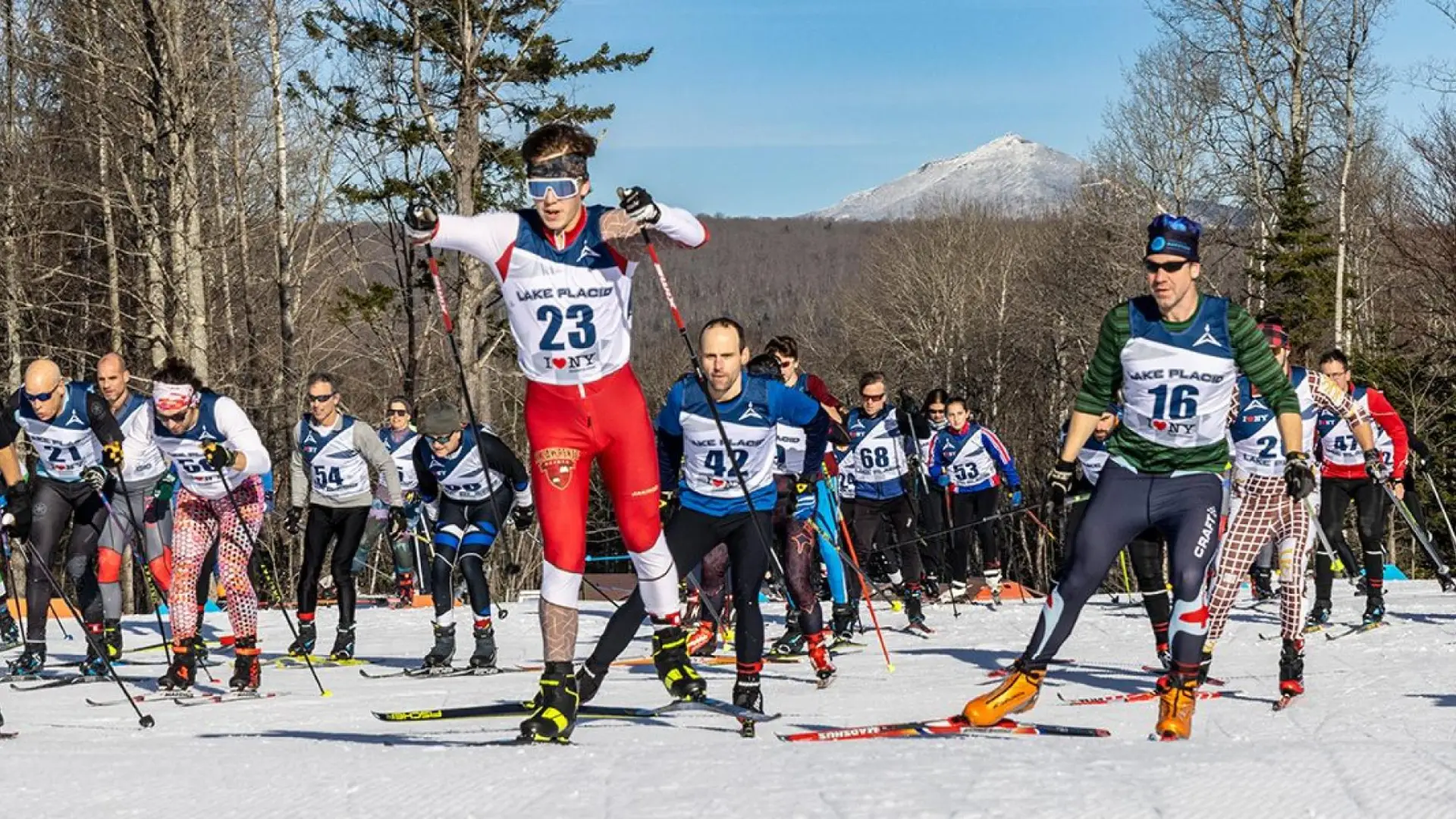 Cross-country skiers in a competitive race on a sunny day, wearing numbered bibs. Snow-covered landscape with bare trees and a distant mountain peak.