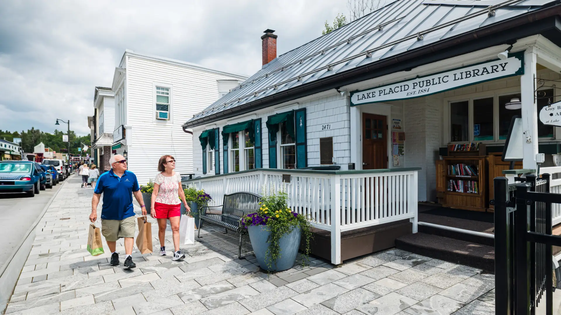Senior couple carrying shopping bags walk past Lake Placid Public Library in summer