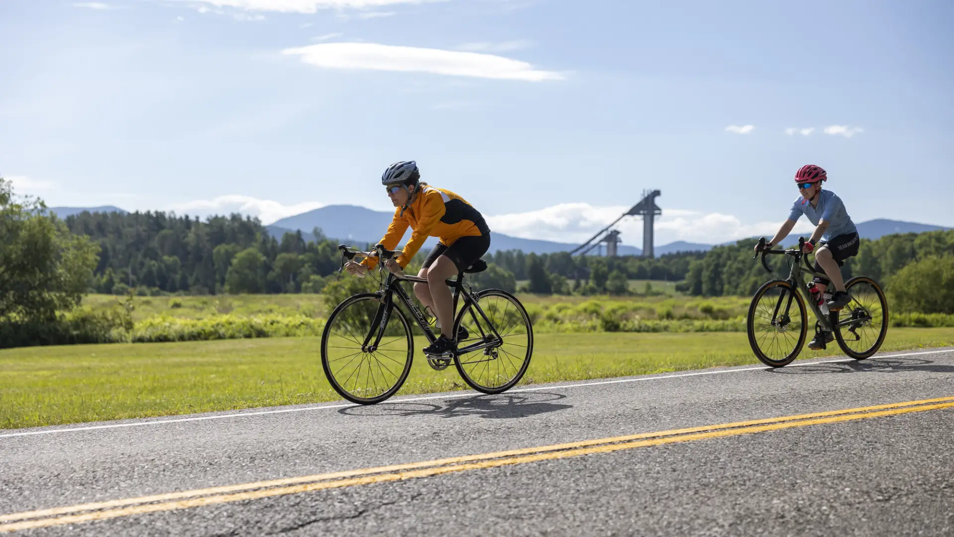 Two cyclists on a road with the Ski Jumps in the background.