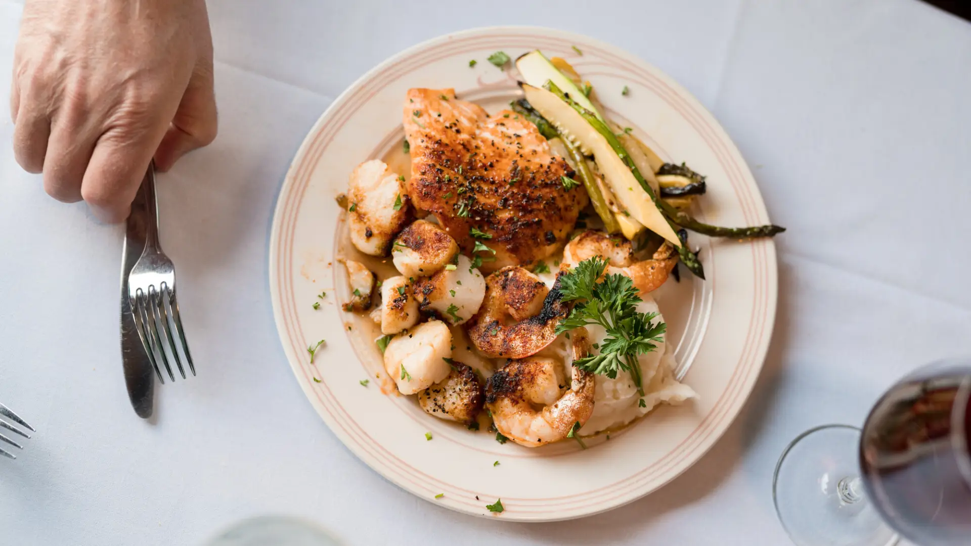 Plate of shrimp, chicken, and vegetables on white tablecloth table with hand holding fork