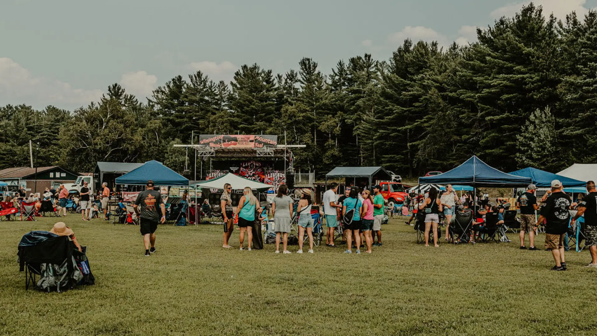 Music festival crowd seen in field with pop-up tents, some people seated in lawn chairs, and some people standing