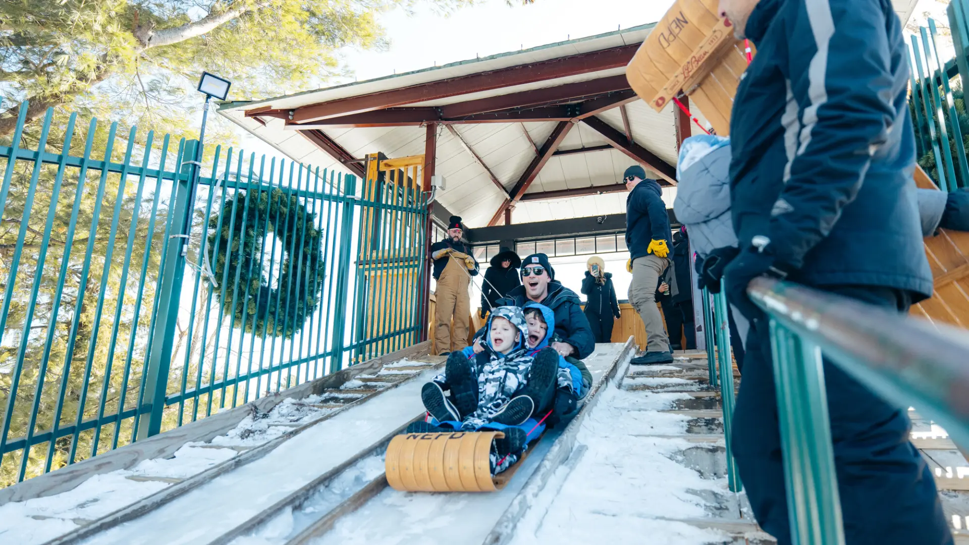 Man sits behind two children on a wooden toboggan, all wearing winter outdoor gear. 
