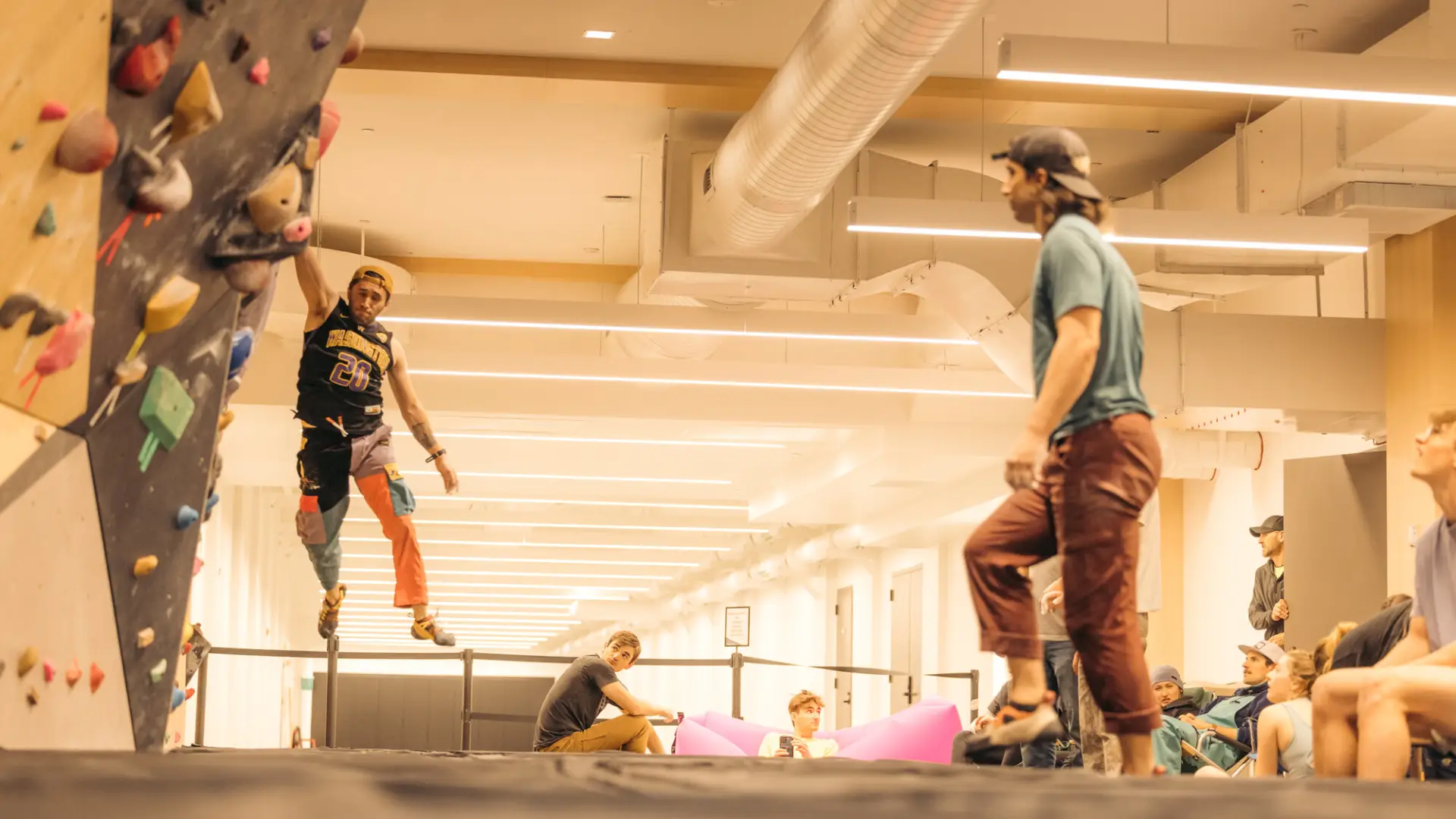 Climber hangs off of bouldering wall by one hand above black mats while other climbers look on