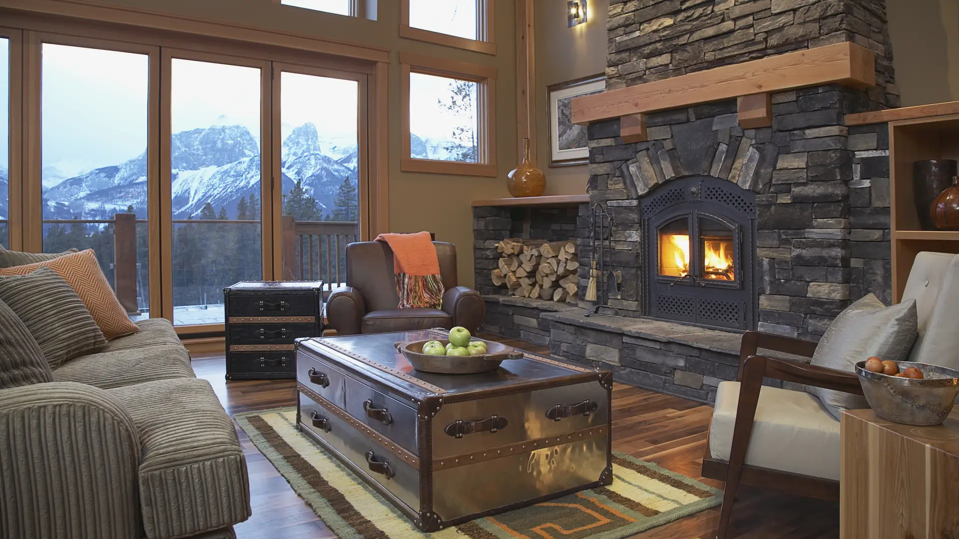 The main living space of a large home with two stories of windows, a stone fireplace with built in wood storage and ample lush seating.