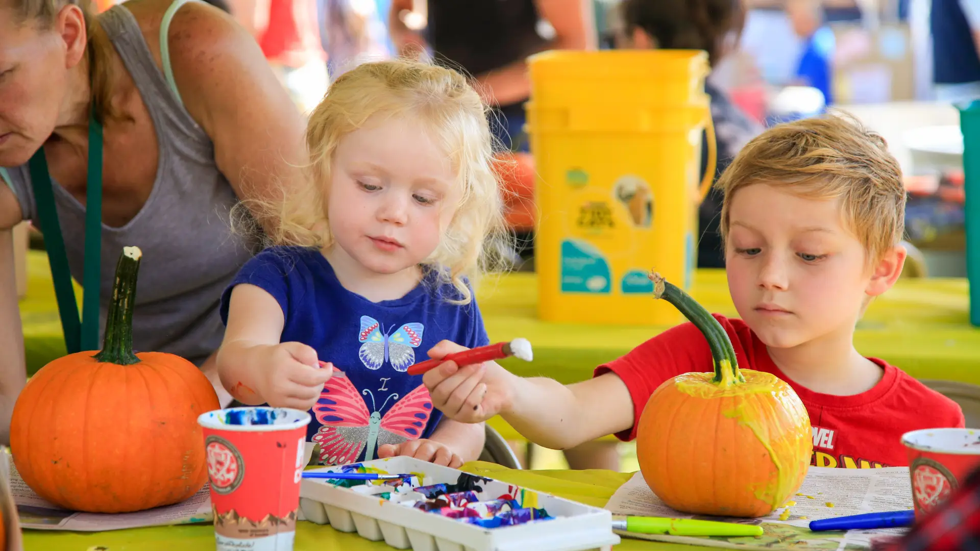 Young girl and young boy sit at table at event painting two small pumpkins