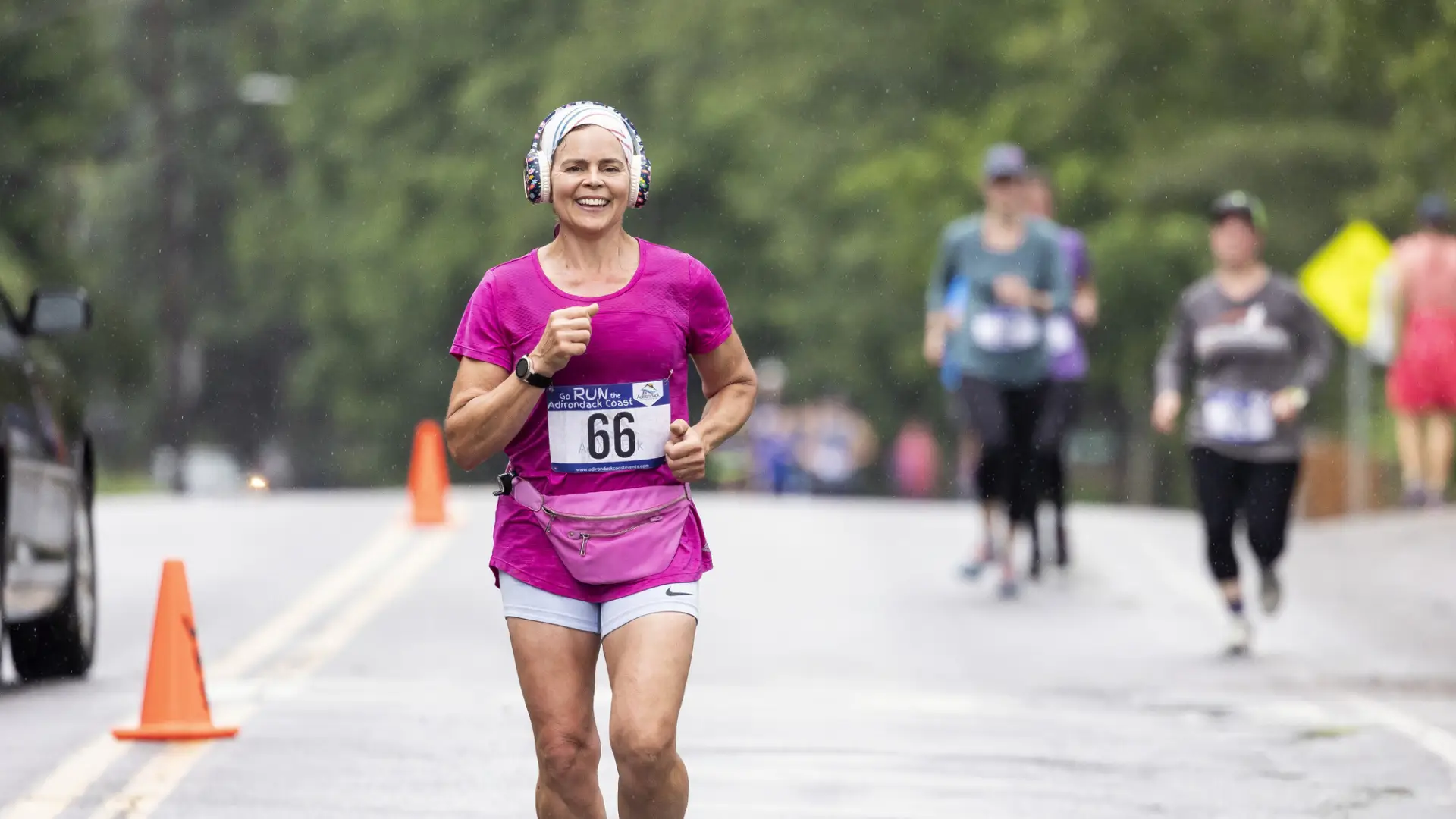 A smiling woman with light skin runs toward the camera during an outdoor race.