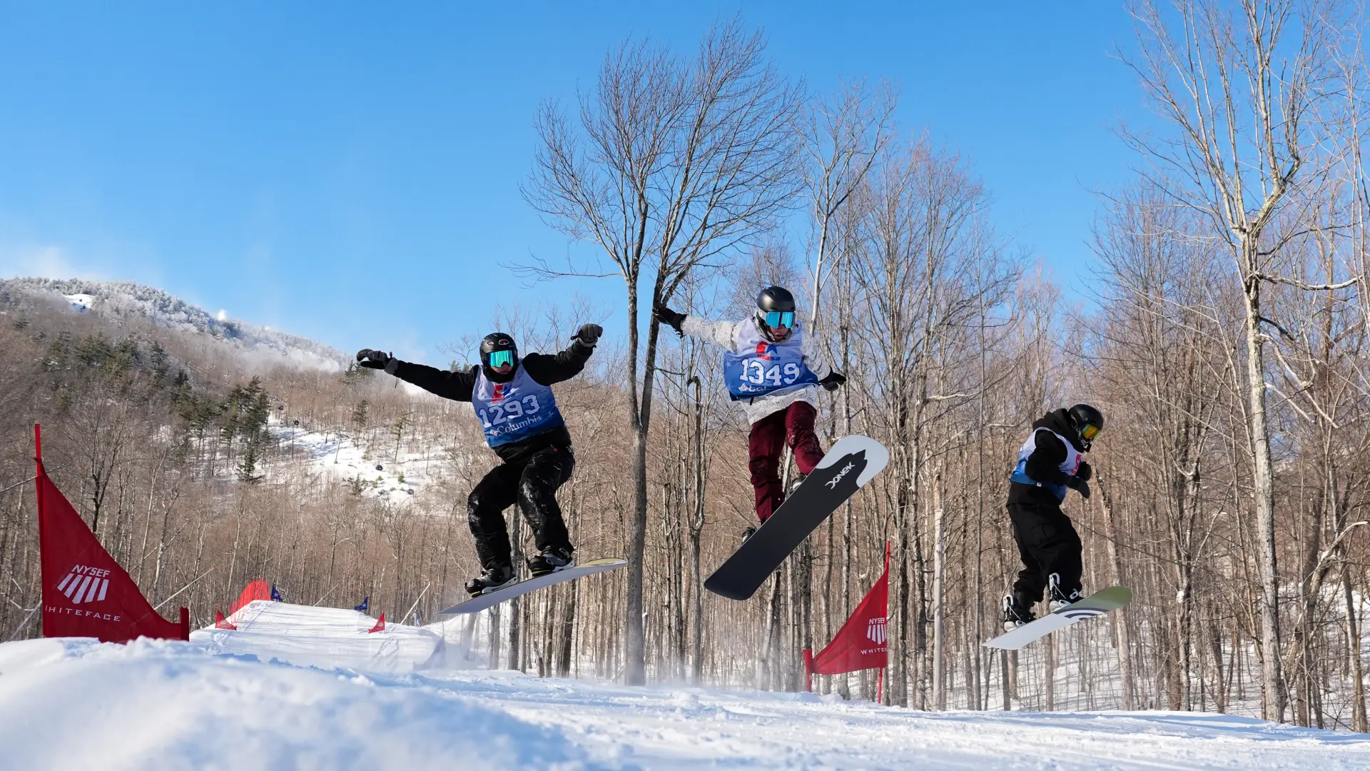 Three snowboarders wearing competition bibs catch air simultaneously during a snowboard cross race at Whiteface Mountain.