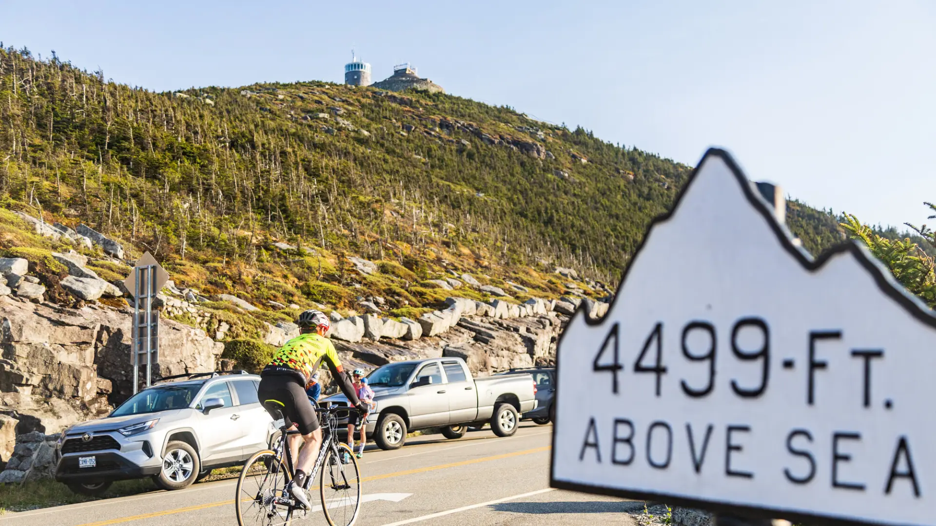 A cyclist rides uphill past parked cars on a mountain road, with a sign in the foreground reading "4499-FT. ABOVE SEA LEVEL".
