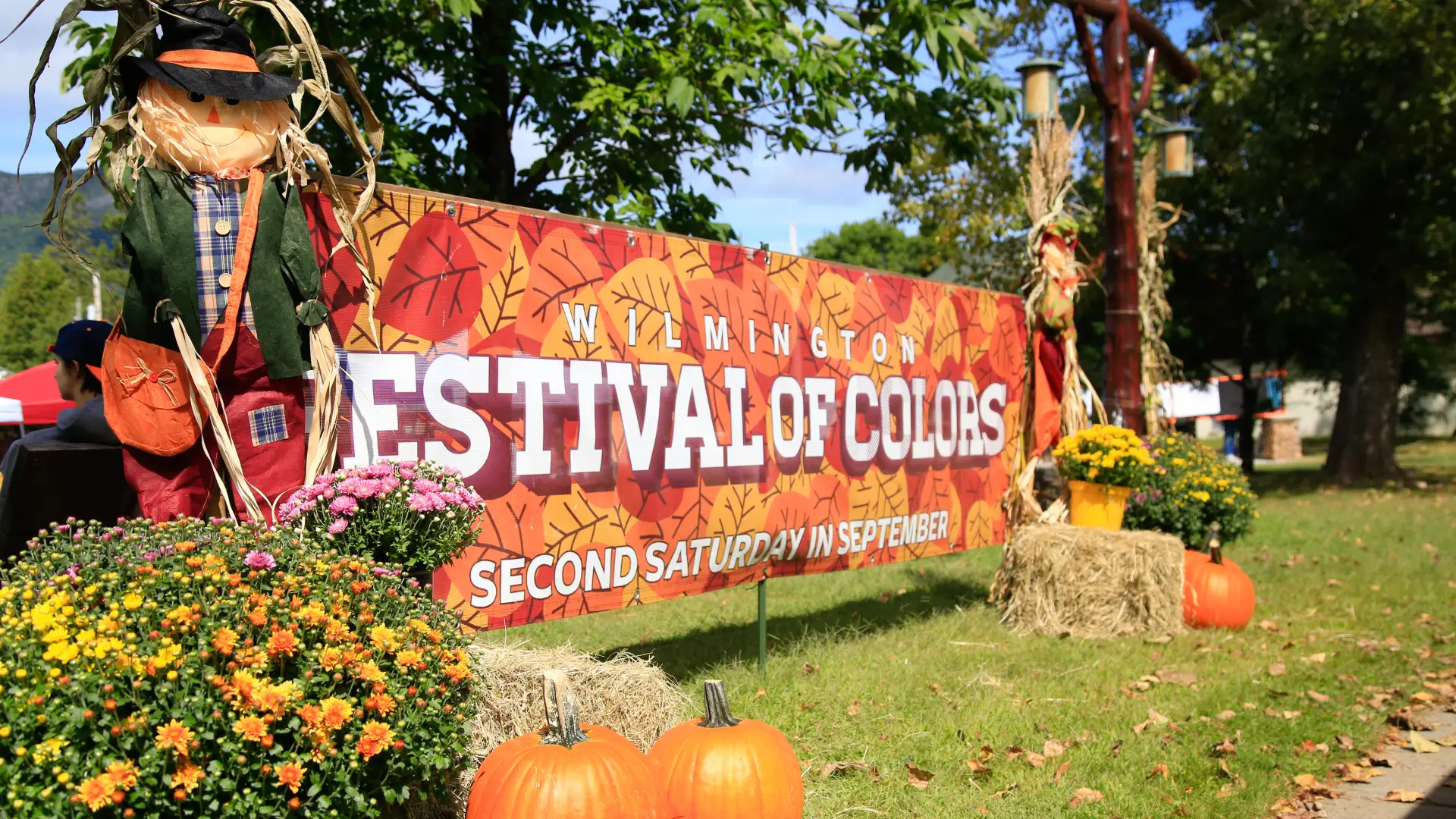 Large Festival of Colors event banner bookended by scarecrows, hay bales, mums, and pumpkins outdoors at park