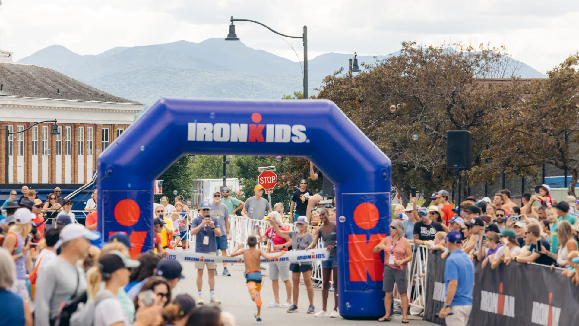 Boy in swim trunks runs toward finish line with blue inflatable arch while spectators cheer at Lake Placid Ironkids race