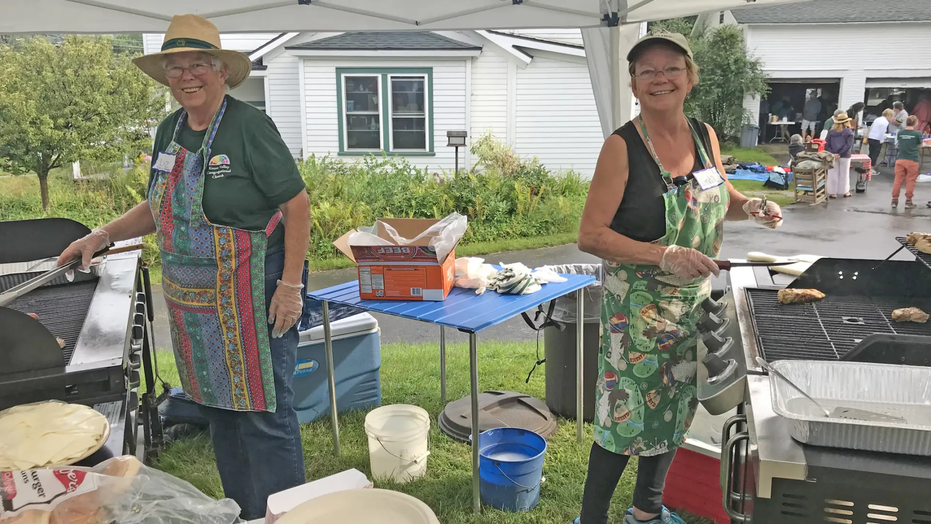 Two senior women cook on grills under pop-up tent at event