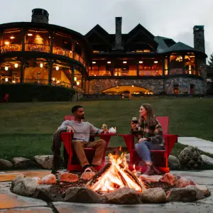 A man and woman sit at a firepit in front of a lodge. 