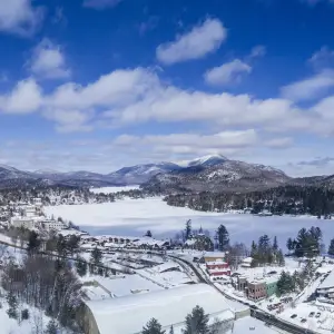 Aerial photo of the Village of Lake Placid in winter. Mirror Lake is covered in snow, as are all the buildings of downtown and the mountains in the distant background. 