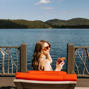 Blonde woman sits in red lounge chair on dock by lake looking right wearing sunglasses and holding cocktail with mountains in the distance