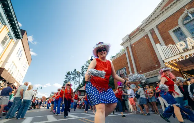 Woman in top hat, red shirt, and blue fringe skirt waves silver pom poms as she dances in a 4th of July parade in Lake Placid