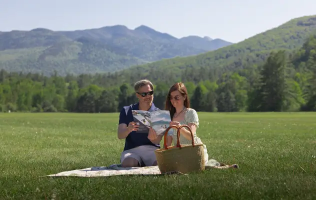 Couple sits on picnic blanket with picnic basket in green field holding Lake Placid Travel Guide with mountains in background